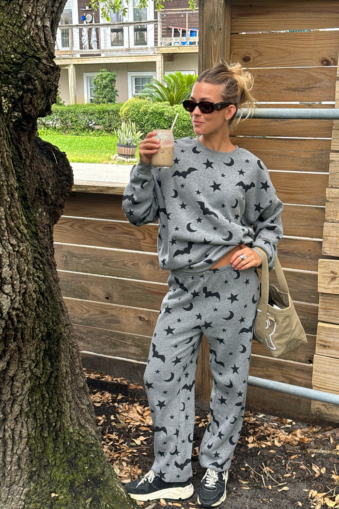 A woman wearing a matching gray lounge set with black crescent moon and star patterns, standing outdoors next to a wooden fence and tree trunk while holding a drink.