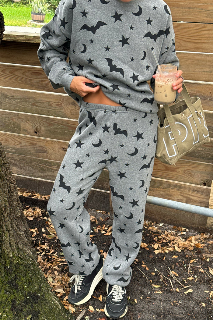 A person wearing a matching gray sweatshirt and jogger sweatpants set with a black bat and moon pattern, standing outdoors in front of a wooden fence and holding a drink and a tote bag.
