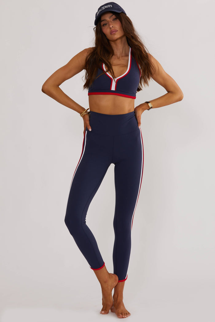 A woman posing in blue and multi-colored active wear leggings with a sports bra and a cap, the outfit features a stars and stripes pattern.