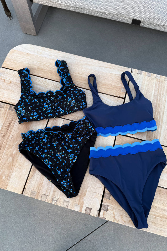 Three-piece navy blue swimwear set arranged on a light wood slatted surface. The set includes a scalloped halter bikini top, matching high-waisted bottoms with a blue floral print, and a matching bandeau-style bikini top with layered scalloped blue ruffles and high-waisted bottoms.