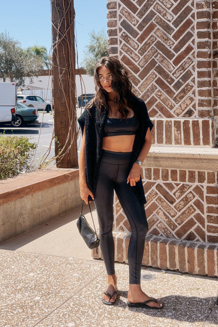 A woman stands outdoors on a sidewalk wearing a black activewear set consisting of a sports bra, high-waisted leggings, and a draped open-front cardigan, paired with black flip-flops and holding a small black handbag.