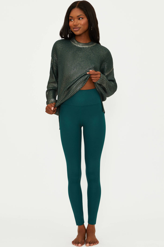 Woman wearing a dark green ribbed sweater and teal leggings on a white background