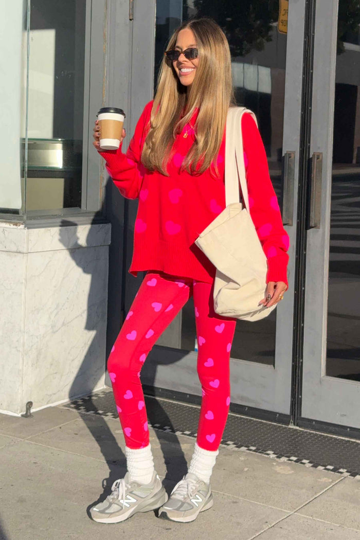 Woman in red sweater and pink heart leggings holding a coffee cup and beige bag outdoors.