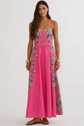 A woman modeling a bright pink maxi dress with intricate multicolor floral embroidery and a matching belt, set against a plain light gray studio background.