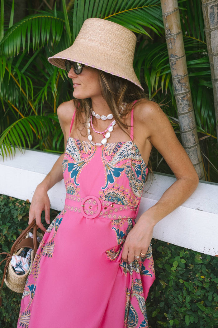 A woman wearing a bright pink sleeveless dress with a multicolor paisley print, a beige straw hat, and sunglasses, posing outdoors against a white fence and green foliage.