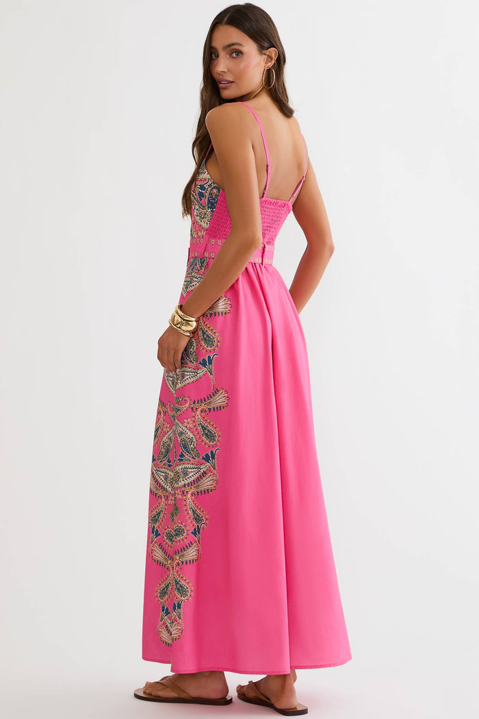 A woman modeling a bright pink maxi dress with intricate multicolor floral embroidery and a matching belt, set against a plain light gray studio background.