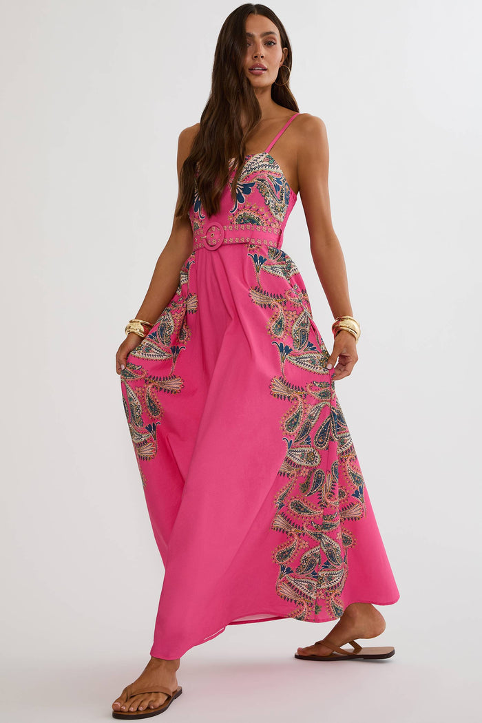 A woman modeling a bright pink maxi dress with intricate multicolor floral embroidery and a matching belt, set against a plain light gray studio background.
