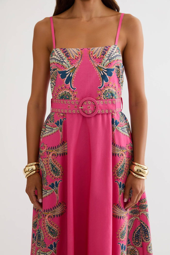A woman wearing a bright pink midi dress with a paisley pattern in blue, gold, and beige, featuring thin straps, a square neckline, and a matching belt with a large round buckle.