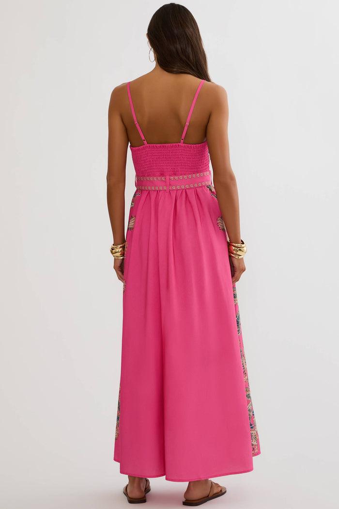 A woman viewed from the back wearing a bright pink sleeveless maxi dress with thin straps, a smocked bodice, and embroidered side panels, against a plain light gray studio background.