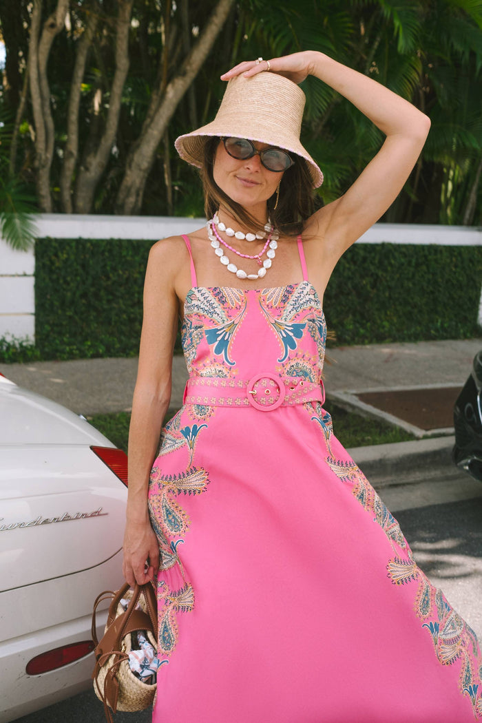 A woman wearing a bright pink sleeveless maxi dress with colorful paisley embroidery, accessorized with a straw hat, sunglasses, and layered pearl necklaces, posing outdoors next to a white car.
