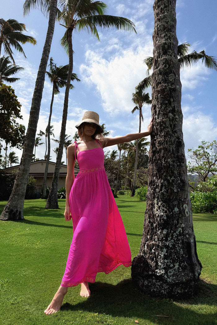 Woman in a pink dress and hat standing next to a palm tree on a grassy area with trees and buildings in the background.