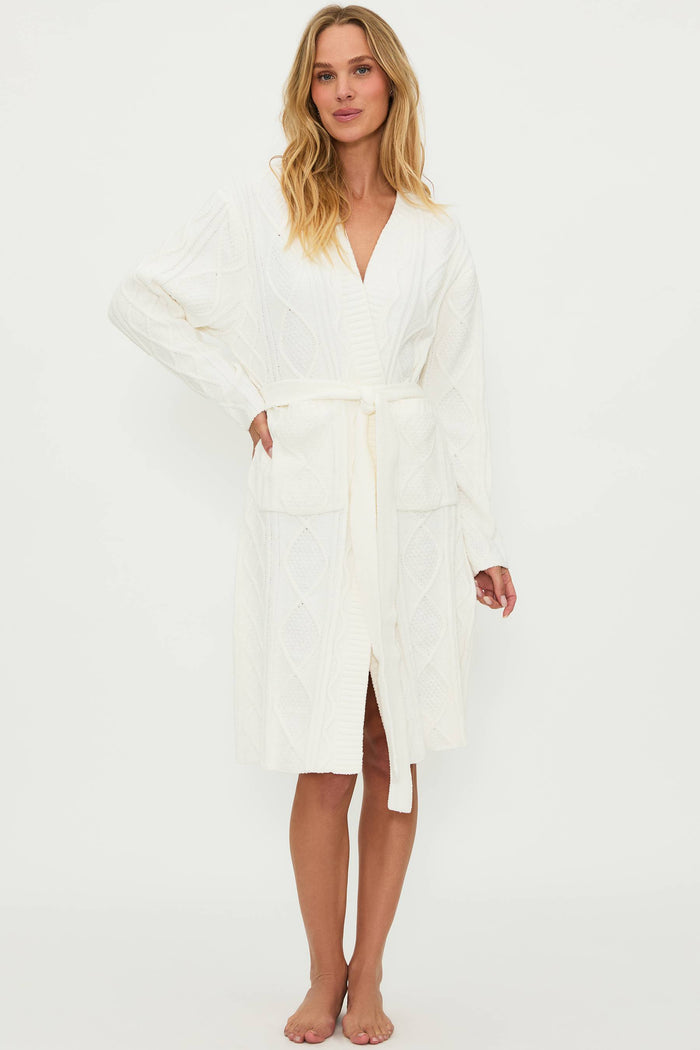 A female model wearing a white cream cable knit robe with a shawl collar and front pockets, belted at the waist.