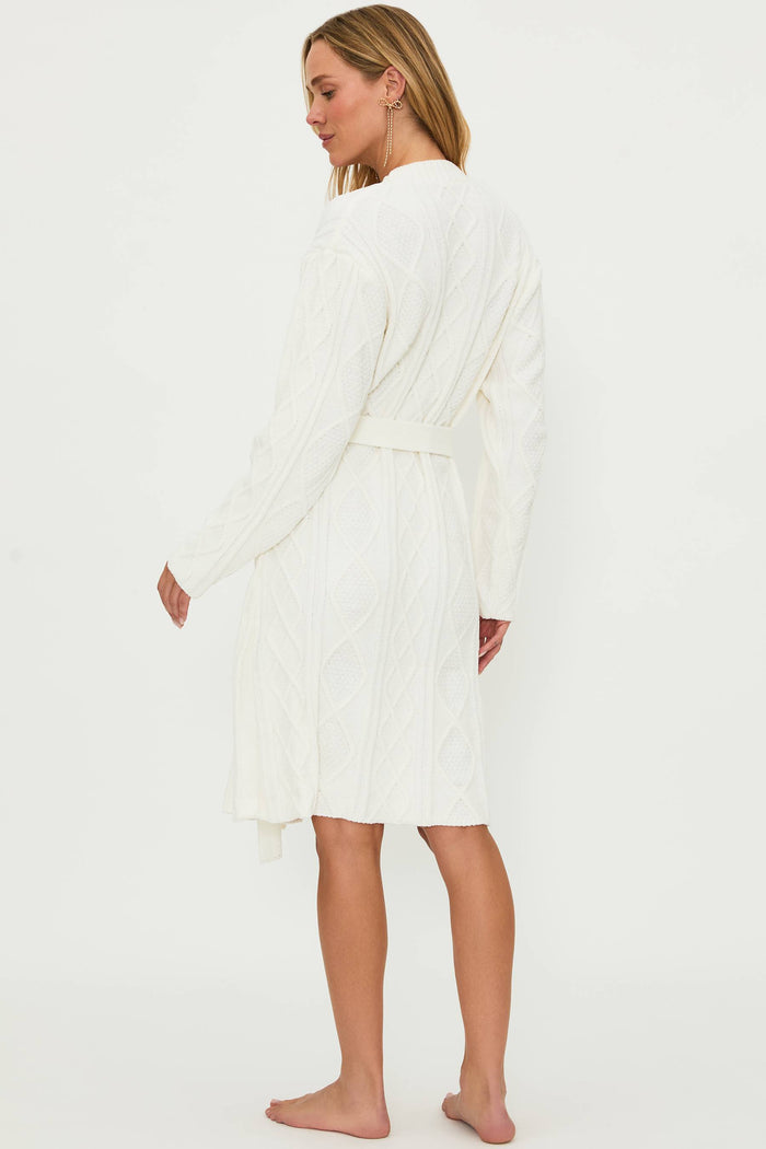 A female model wearing a white cream cable knit robe with a shawl collar and front pockets, belted at the waist.