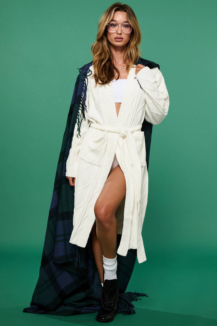 A female model wearing a white cream cable knit robe with a shawl collar and front pockets, belted at the waist.