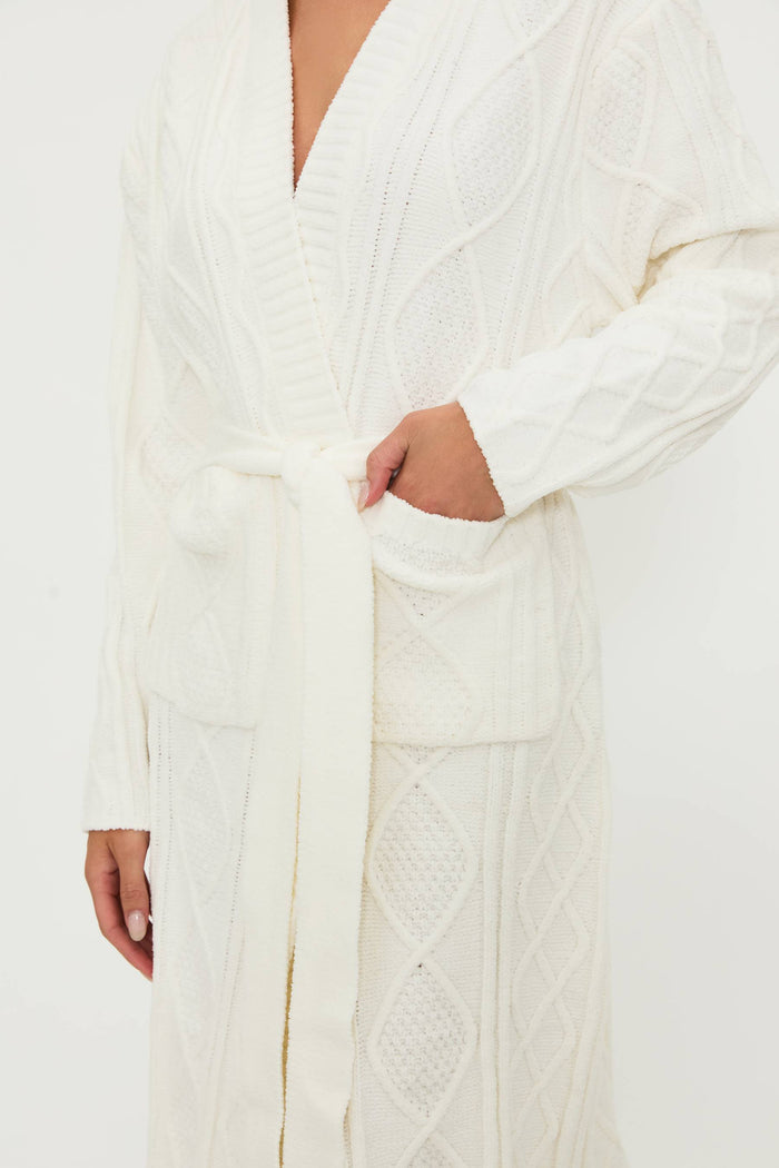 A female model wearing a white cream cable knit robe with a shawl collar and front pockets, belted at the waist.