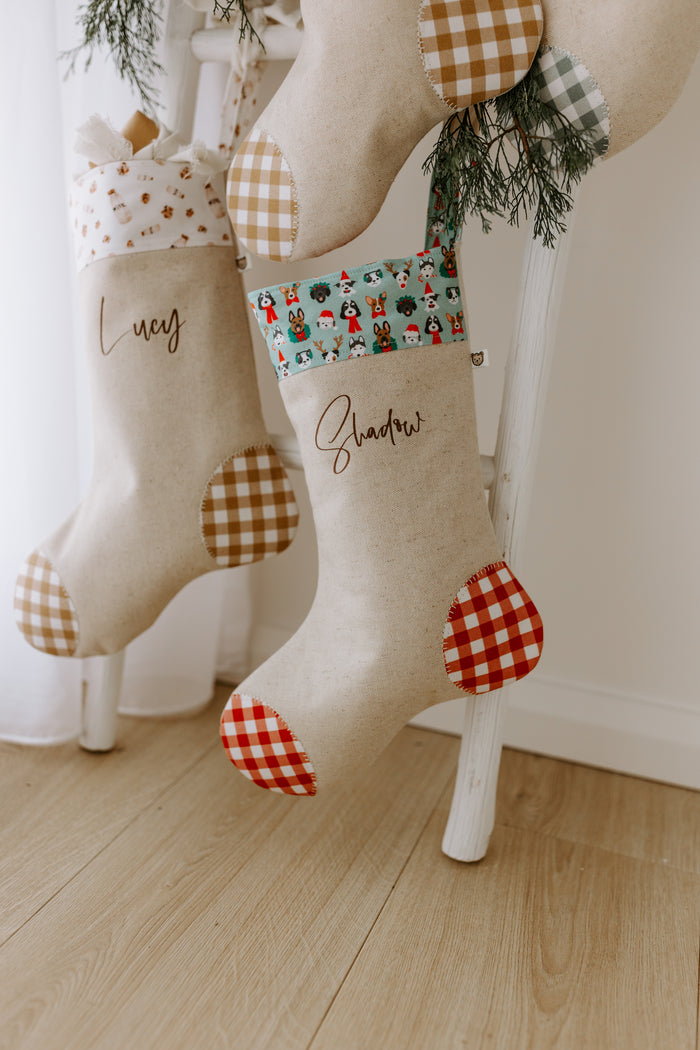 Pair of personalised Christmas stockings hanging on a white wooden ladder. One stocking reads “Lucy” with a neutral gingham trim, while the other says “Shadow” with a festive cuff featuring colourful cartoon dog faces in Santa hats, scarves, and wreaths. Both stockings are crafted from natural linen with red or tan gingham heel and toe patches. A perfect Christmas decoration for pet lovers, fur babies, and families wanting to include their dogs in holiday traditions.