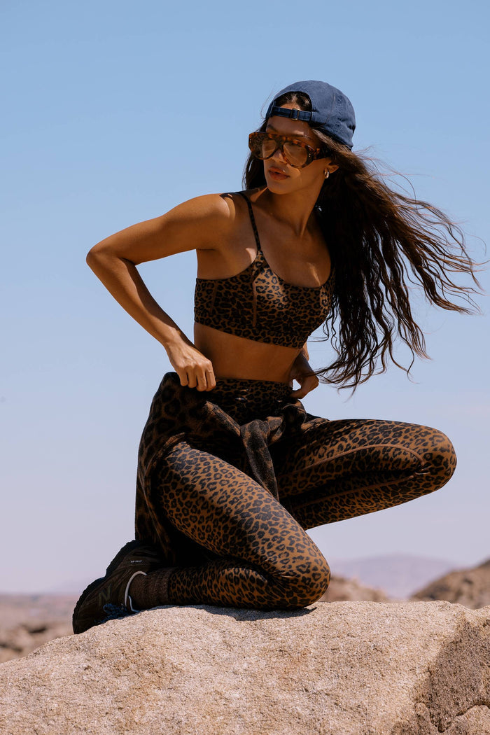 A woman wearing a matching leopard-print activewear set consisting of a sports bra and leggings, posing on a large rock outdoors under a clear blue sky.