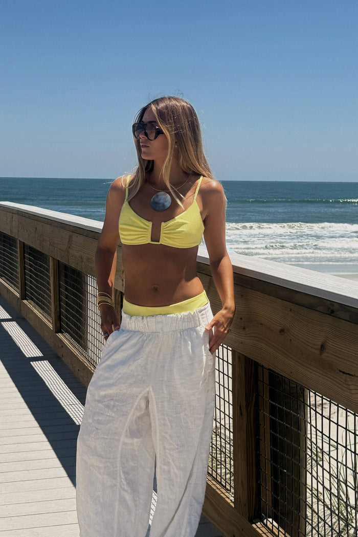 A woman stands on a wooden boardwalk by the ocean wearing a bright yellow bikini top and white wide-leg pants.