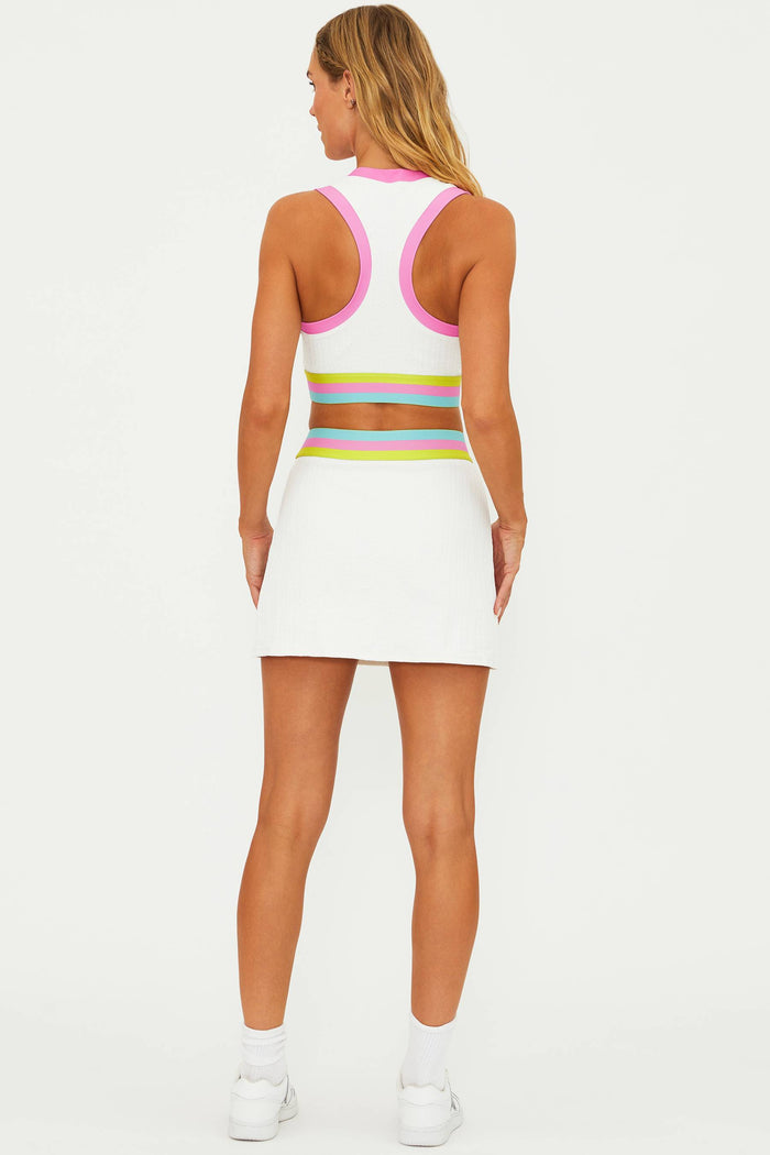 A woman wearing a white tennis skirt with a colorblock waistband in pink and yellow tones, paired with a matching white tank top, and white tennis shoes.