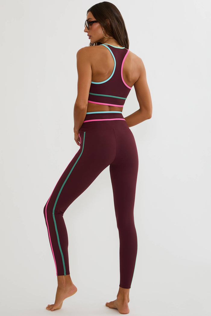 A woman modeling a matching athletic set consisting of a racerback sports bra and high-waisted leggings in deep burgundy with neon pink and mint green stripe accents, shown from a rear three-quarter view against a plain white studio background.