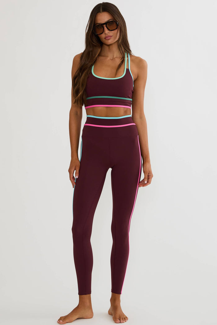 A woman modeling a matching burgundy activewear set consisting of a sports bra and high-waisted leggings with thin multicolor stripe accents in mint green, pink, and teal, photographed against a light gray studio background.
