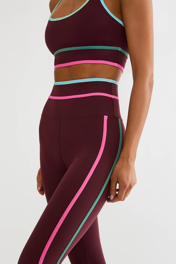 A close-up of a model wearing a matching athletic set in deep burgundy with bright pink and mint green contrast trim, including a sports bra and high-waisted leggings.