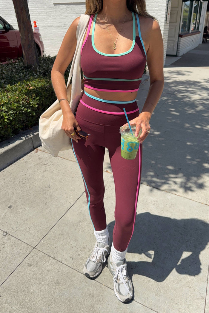 A woman wearing a matching athletic set consisting of a maroon sports bra and leggings with light blue and pink trim, standing on a sunny sidewalk.