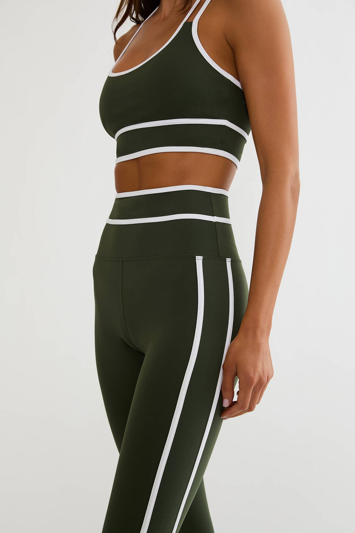 A woman wearing a matching dark olive green activewear set with white contrast trim, including a sports bra and high-waisted leggings, shown against a plain light gray studio background.