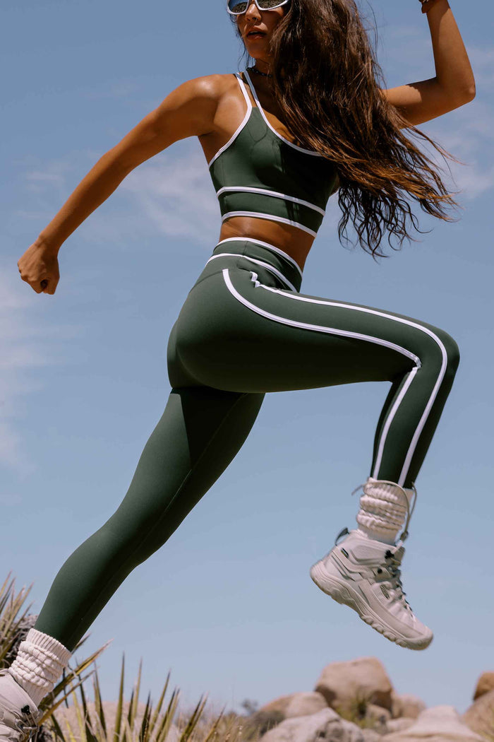A woman in a dark green and white athletic set jumps outdoors against a blue sky, wearing a matching sports bra and leggings with white side stripes.
