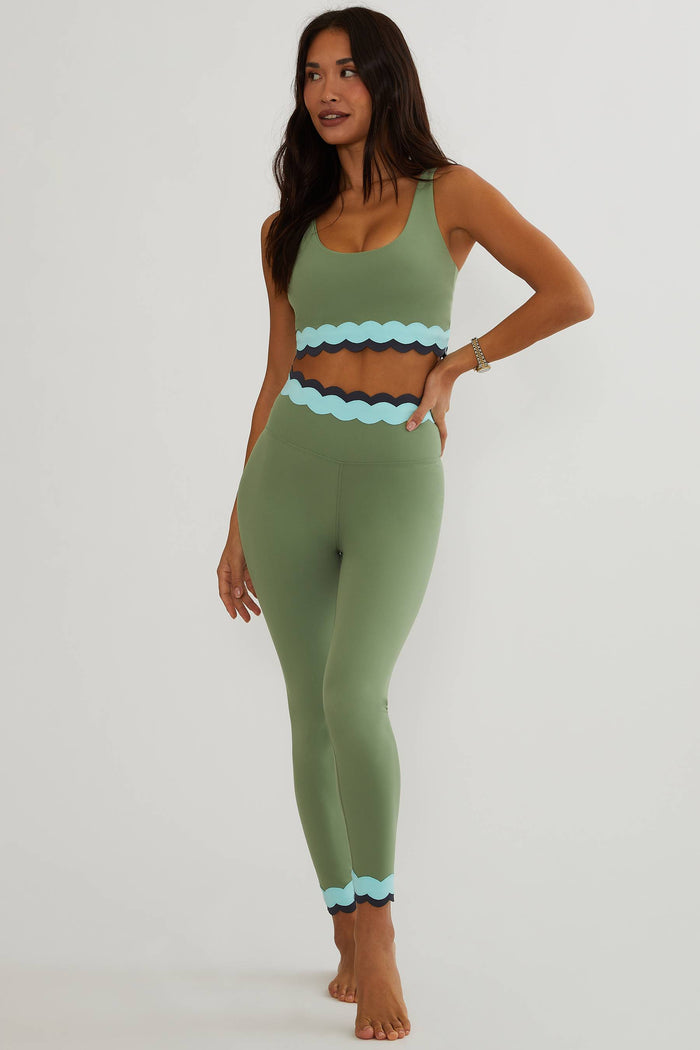 A woman modeling a matching sage green activewear set with a scoop-neck tank top and high-waisted leggings, both featuring a light blue scalloped trim at the waist and hem, against a plain light gray studio background.