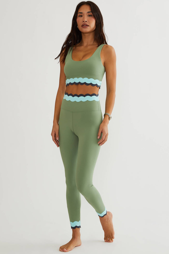 A woman modeling a matching sage green activewear set consisting of a scoop-neck crop top and high-waisted leggings, featuring a wavy light blue and dark brown stripe pattern at the waist and hem.