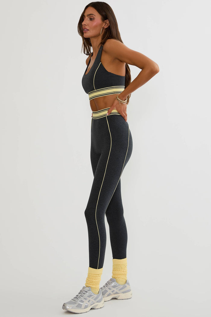 A woman modeling a matching dark gray activewear set consisting of a sports bra and high-waisted leggings with yellow and white stripe accents, paired with yellow socks and gray sneakers, against a light gray studio background.