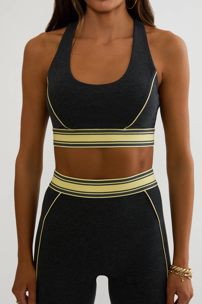A woman wearing a dark charcoal gray athletic set with light yellow stripes. The set includes a halter-neck sports bra and matching high-waisted leggings with contrast yellow piping and a wide waistband.