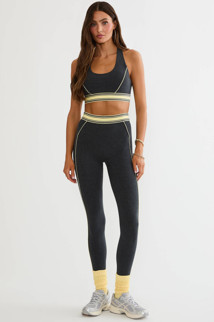 A woman modeling a matching dark gray athletic set consisting of a sports bra and high-waisted leggings, both featuring a horizontal stripe pattern in yellow, white, and gray. She is also wearing yellow socks and gray sneakers against a plain white studio background.