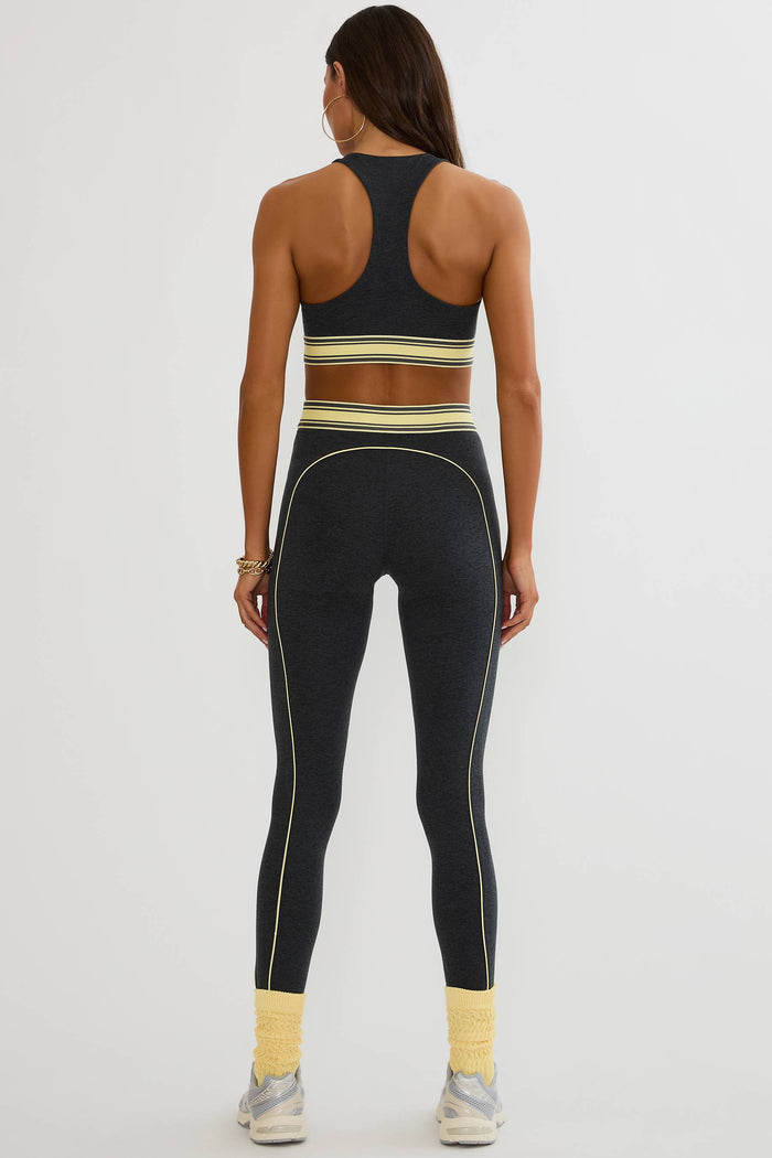 A woman shown from the back wearing a matching black activewear set with thin yellow stripes and piping details, paired with yellow socks and white sneakers, against a light gray studio background.