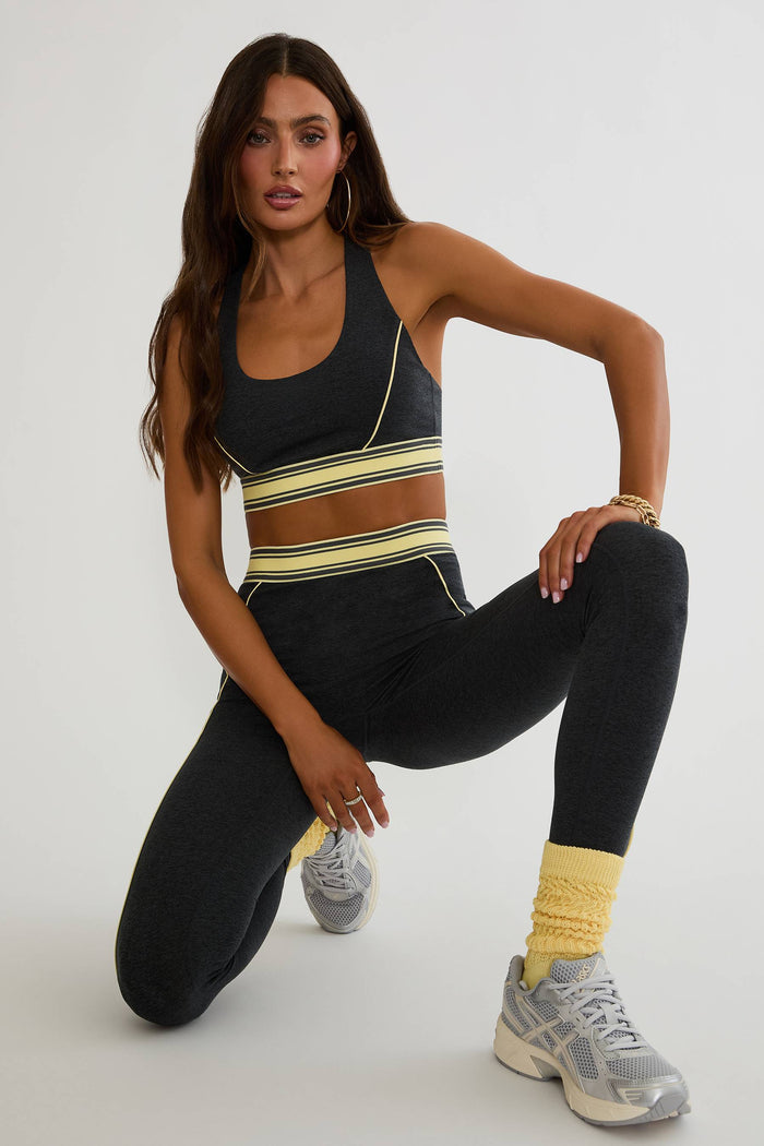 A woman posing in a black athletic set consisting of a sports bra with horizontal yellow and white stripes, matching black leggings, yellow leg warmers, and gray sneakers against a light gray studio background.