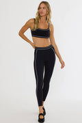 A woman modeling a matching black activewear set with white stripe trim, consisting of a sports bra and high-waisted leggings, against a plain white studio background.