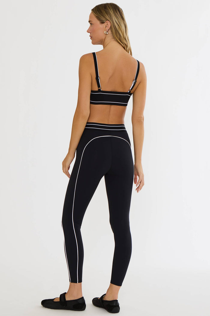 A woman shown from the back wearing a matching black activewear set with white contrast piping, consisting of a sports bra and high-waisted leggings, against a plain white studio background.