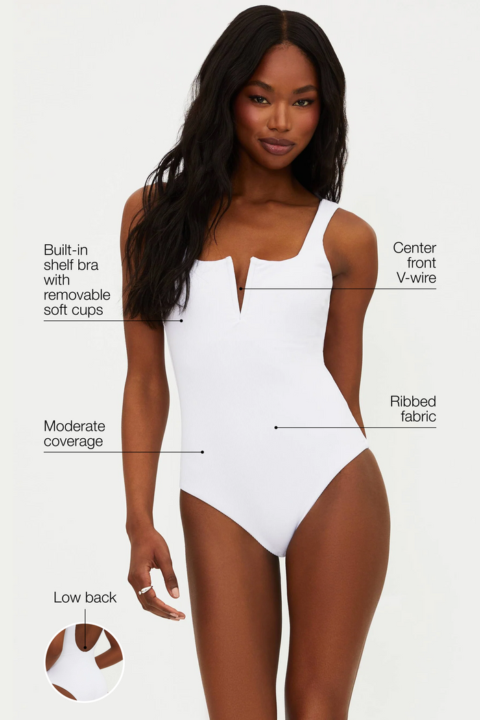 A model is wearing a white one-piece swimsuit with a ribbed texture, fixed straps, and a low back.