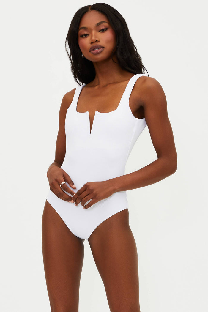 A model is wearing a white one-piece swimsuit with a ribbed texture, fixed straps, and a low back.