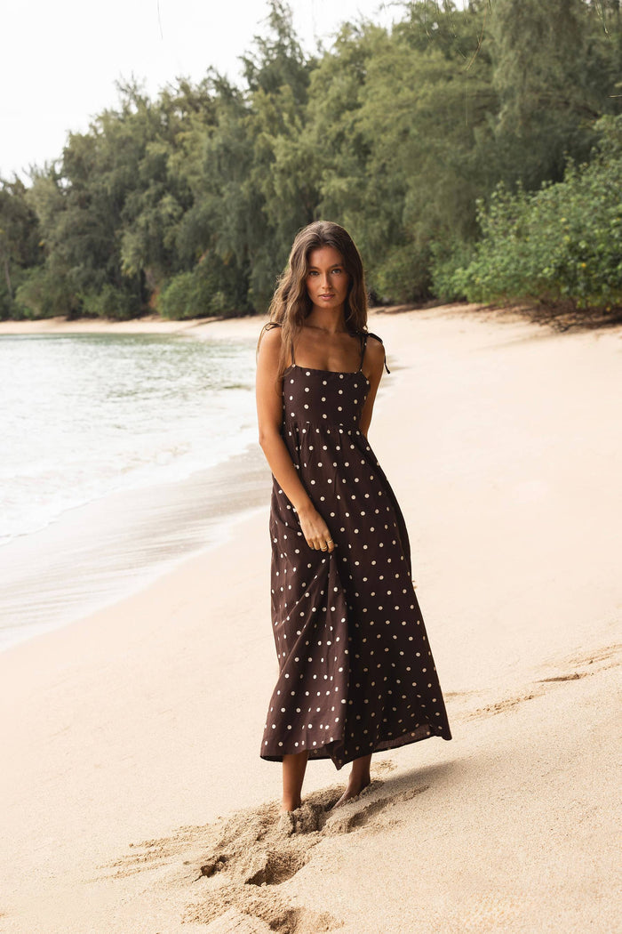 Woman in a polka dot dress standing on a sandy beach with trees in the background