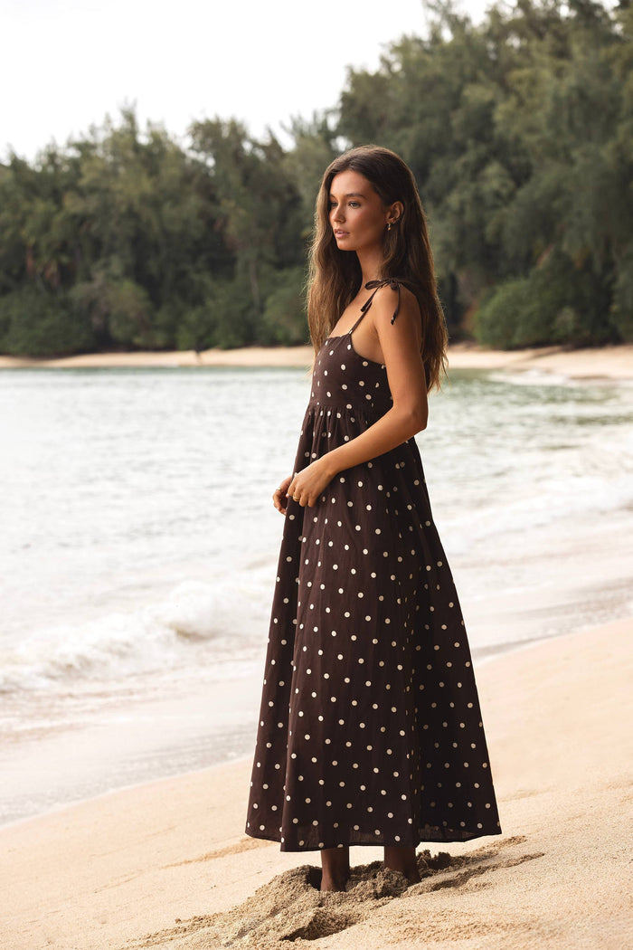 Woman in a polka dot dress standing on a beach with water and trees in the background