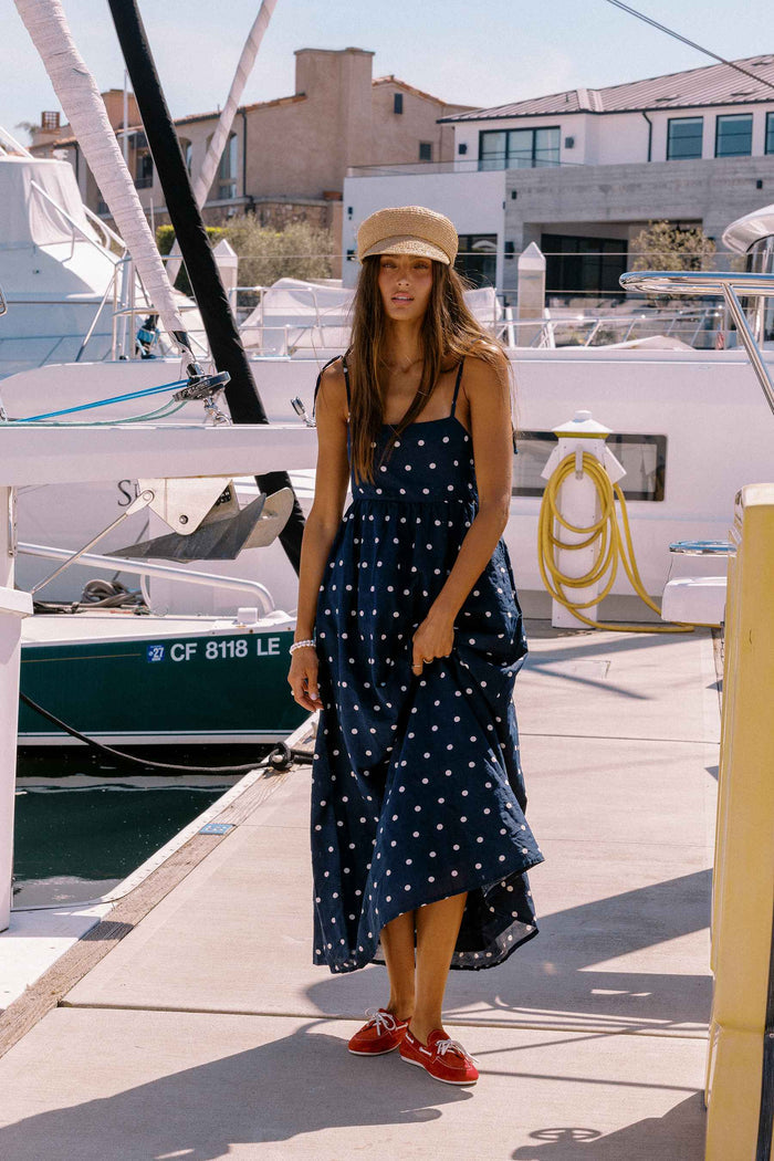 Woman in a navy blue dress with white polka dots and a beige hat standing on a dock with boats in the background.