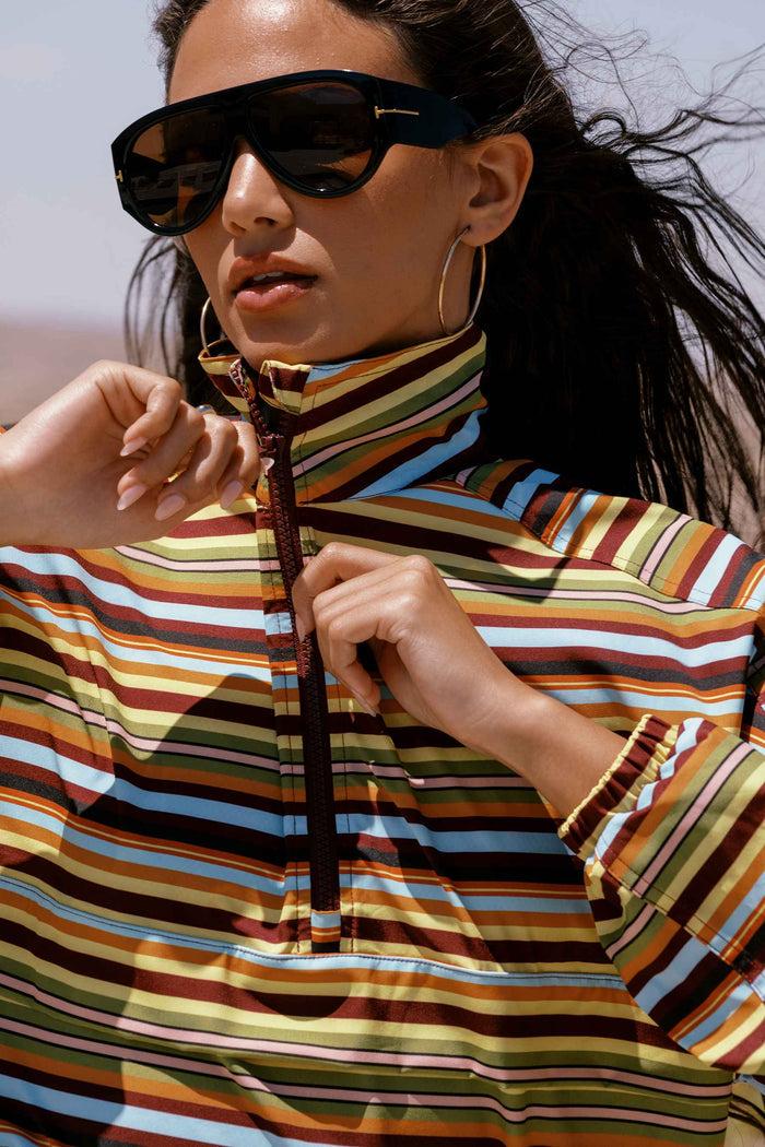 A close-up lifestyle shot of a model wearing oversized black sunglasses, gold hoop earrings, and a colorful horizontally striped high-collar top with a half-zip front. The background is a bright, sunlit outdoor sky.