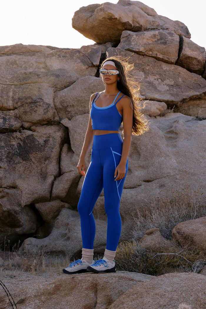 A woman wearing a matching blue athletic crop top and leggings set with white trim stands outdoors in front of large rock formations, wearing white sneakers and white-framed sunglasses.