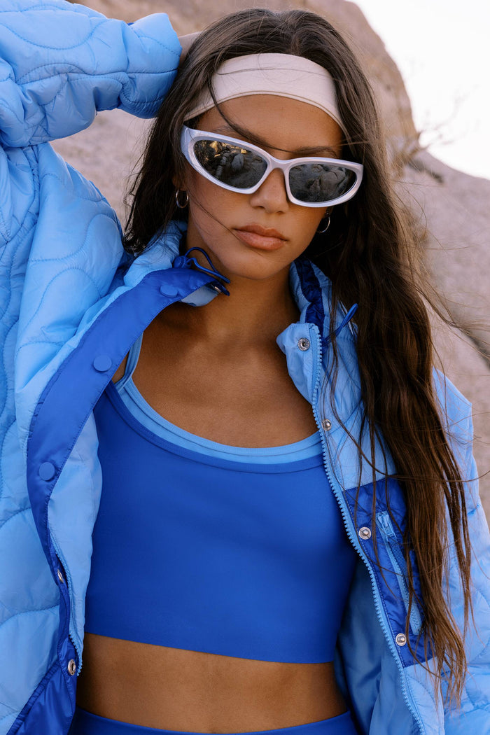 A woman wearing a bright blue puffer jacket over a matching blue sports bra and shorts, with white-framed sunglasses and a beige headband, posed outdoors against a rocky background.