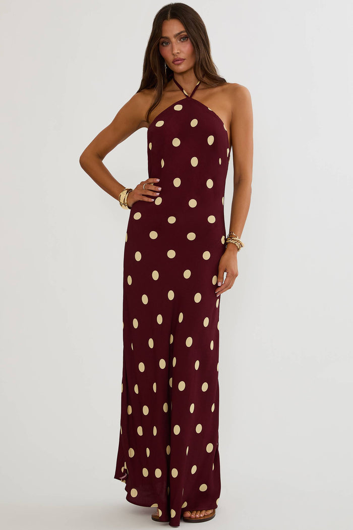 A woman modeling a halter-neck maxi dress in burgundy with a large beige polka dot pattern, set against a plain light gray studio background.