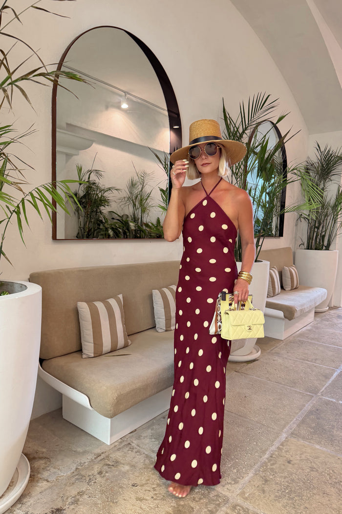 Woman in a polka dot dress and hat standing in a stylish interior setting with plants and furniture.