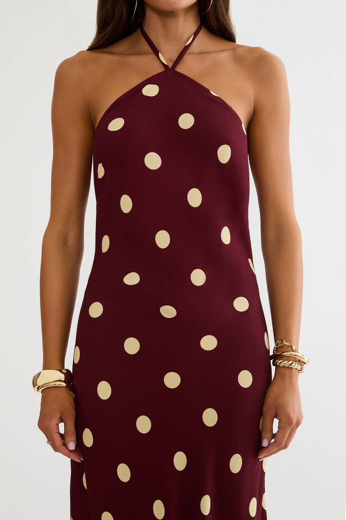A woman wearing a halter-neck dress in burgundy with large cream polka dots, shown from the neck to the upper thighs against a plain light background.