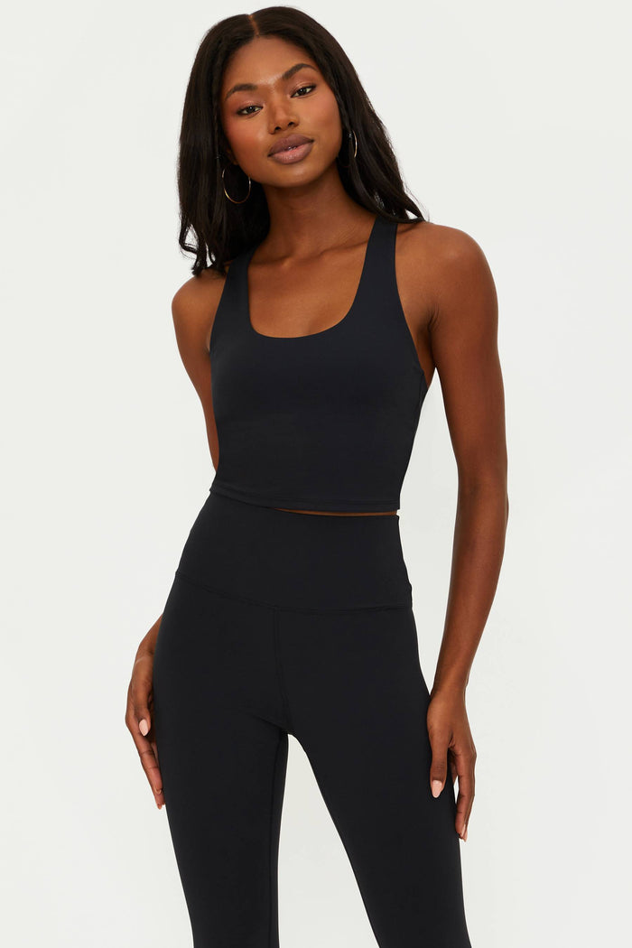 A female model wearing a black matte longline crop top with a scoop neckline and a racerback design.